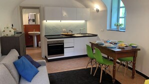 A compact kitchen with a central island, a dining area with a wooden table and green chairs, and a small window with a plant. - 1 Bed Cottage Sleeps 4 Private Garden - Pool (Colletta)