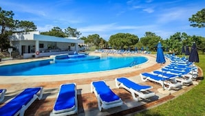 A swimming pool with blue lounge chairs and umbrellas, surrounded by trees and a building. - Scarlet's Place - Spacious 1 bed Quinta Do lago (Quinta do Lago)