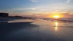 Ein Strand mit Sonnenuntergang, ruhigen Wellen und einem Gebäude im Hintergrund.