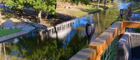 A riverside deck with wooden railings and wicker furniture, overlooking a calm river and lush greenery.