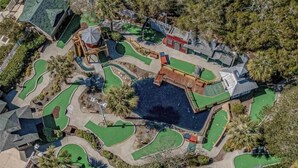 Eine Vogelperspektive auf einen Golfplatz mit mehreren Grünflächen, einem Wasserbecken in der Mitte und umliegenden Bäumen.