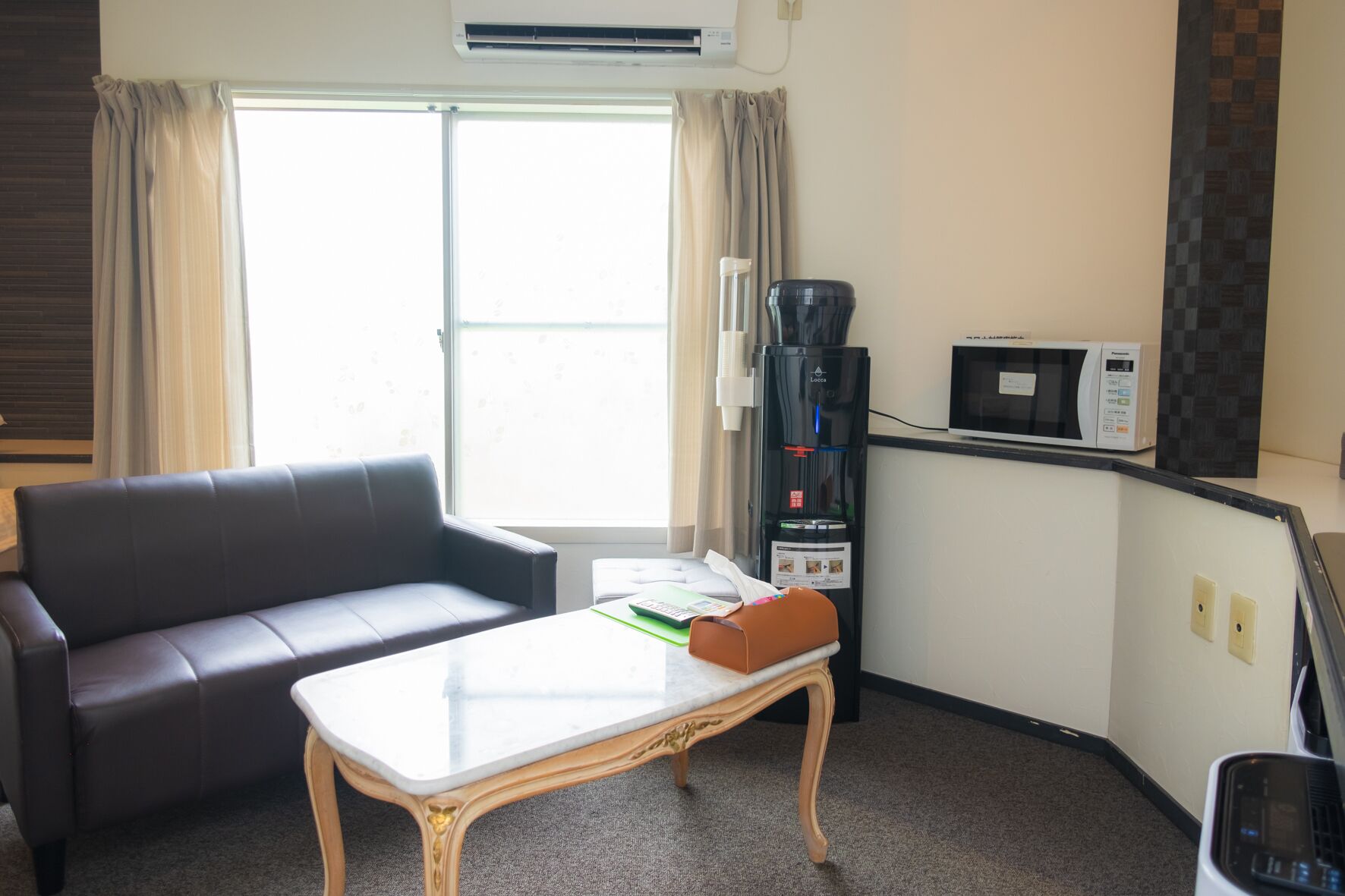 A hotel room with a leather sofa, a small table, a coffee machine, a microwave, and a window with curtains.