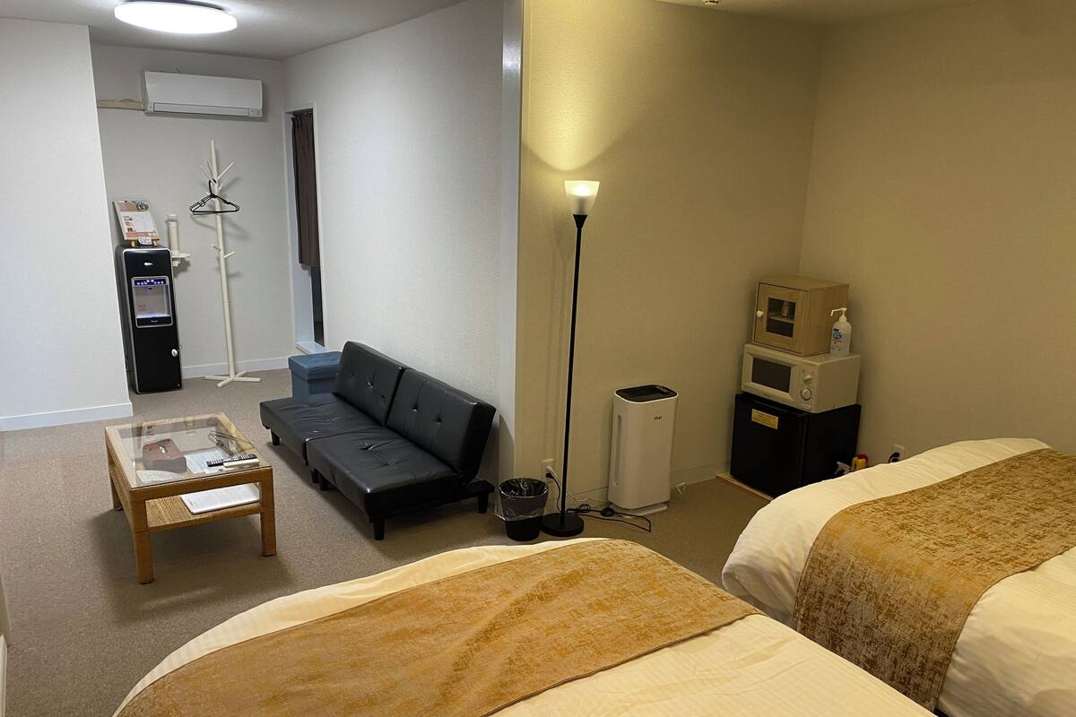 A hotel room with two beds, a sofa, a small table, a lamp, a microwave, and a trash bin.