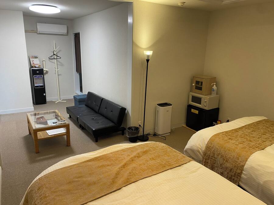 Habitación de hotel con dos camas, un sofá, una mesita, una lámpara, microondas y un cubo de basura.