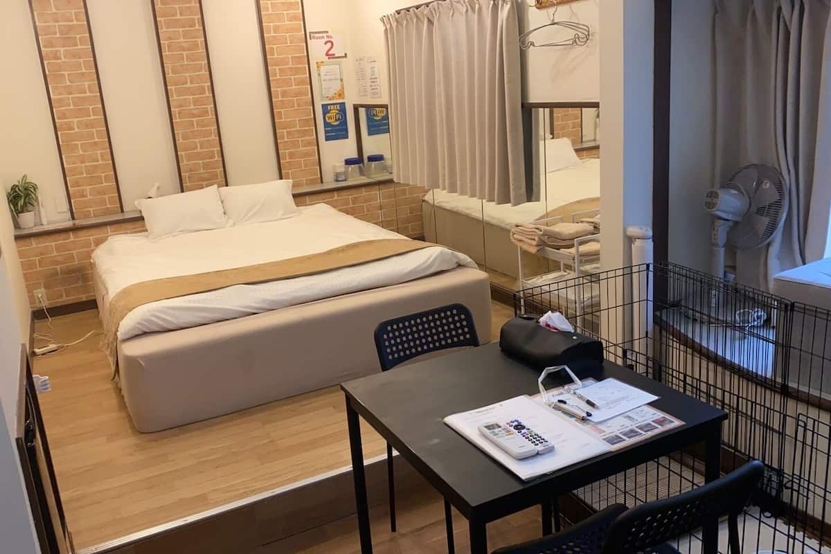 A compact hotel room with a bed, a small table with a calculator and papers, a chair, a window with curtains, and a brick wall.