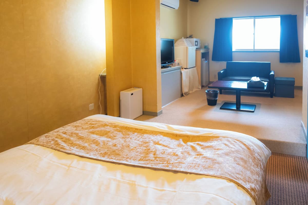 A hotel room with a bed, a desk, a chair, a window, and a kitchenette.