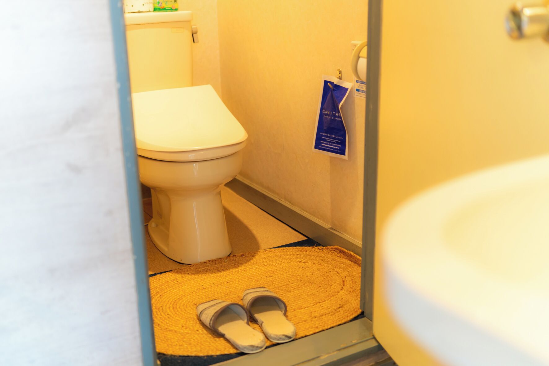 Et badeværelse med toilet, en blå håndspritdispenser og et par slippers på en vævet måtte.