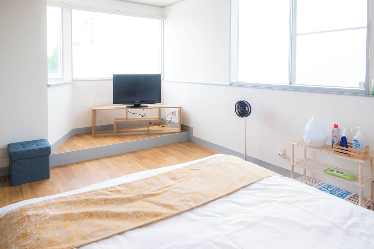 A modern hotel room with a bed, a television on a wooden stand, a standing fan, and a small shelf with toiletries.