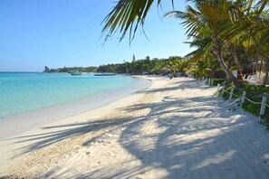 A sandy beach with clear turquoise water, palm trees, and a distant shoreline with buildings. - Mission Gate 4C 2-bedroom condo by Island House Vacation Rentals (Roatan)