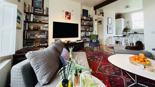 A living room with a sofa, a coffee table, and a TV. There are bookshelves and a dining area with chairs and a table.