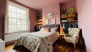 A bedroom with a large window, a bed with floral bedding, a chair, and a wardrobe with hanging clothes.