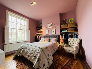 Quarto com uma janela grande, cama com roupa de cama floral, uma cadeira e um guarda-roupa com roupas penduradas.