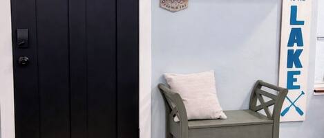 Une porte noire avec des panneaux en verre dépoli, un banc avec un coussin, une cloche fixée au mur, une étagère avec des objets décoratifs et une pancarte sur laquelle on peut lire : « BIENVENUE AU BORD DU LAC ».