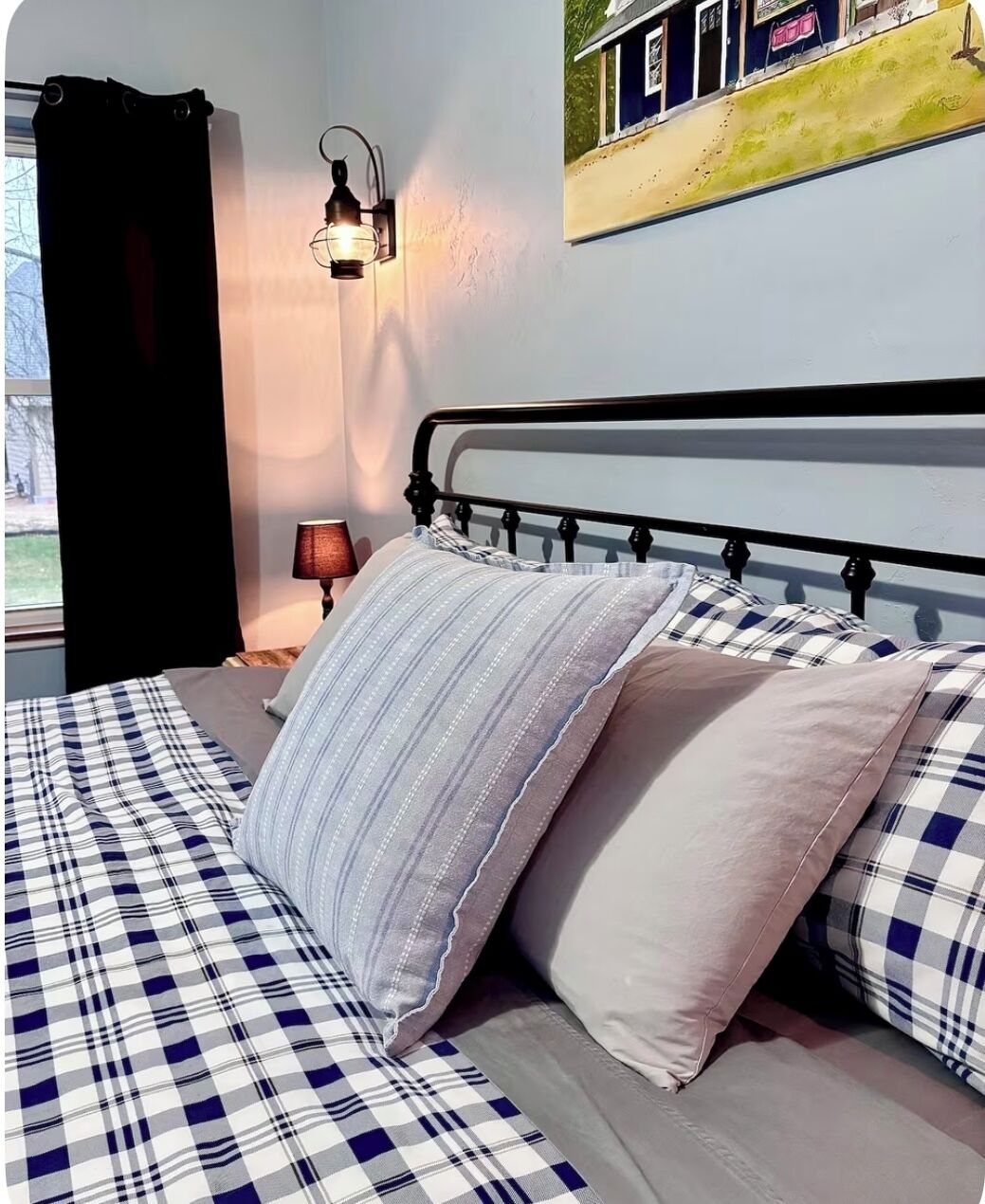 Uma cama com colcha xadrez, duas almofadas, um abajur fixado na parede e um quadro de uma casa.