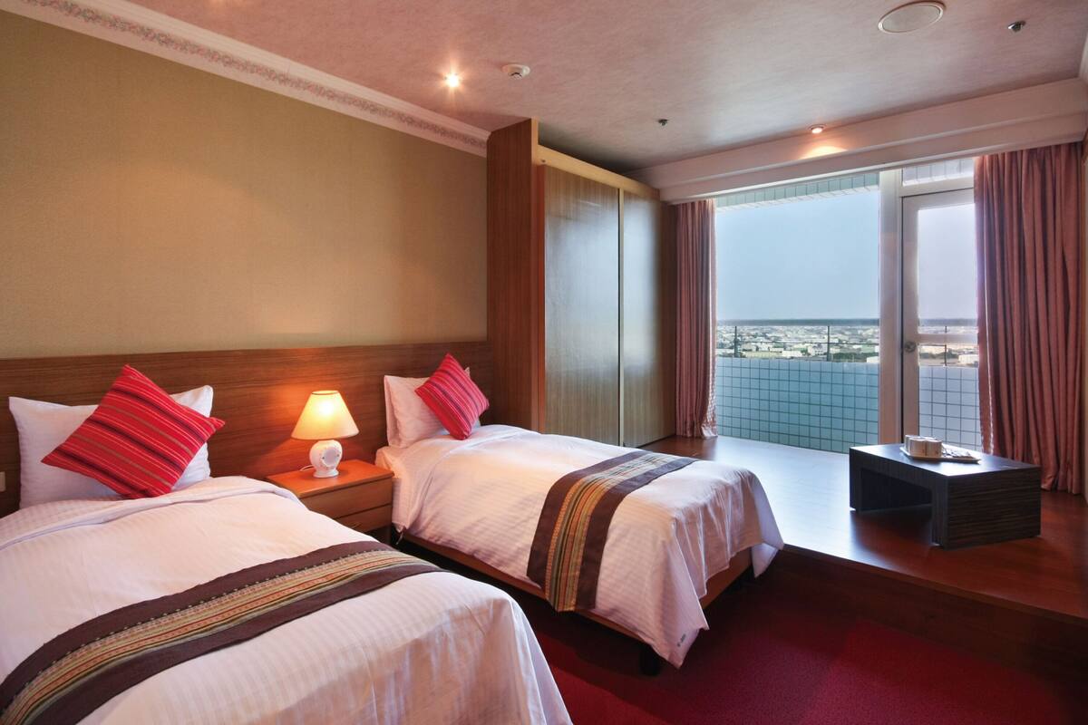 Quarto de hotel com duas camas, uma mesa de cabeceira com abajur, uma mesinha e uma janela grande com vista para a cidade.