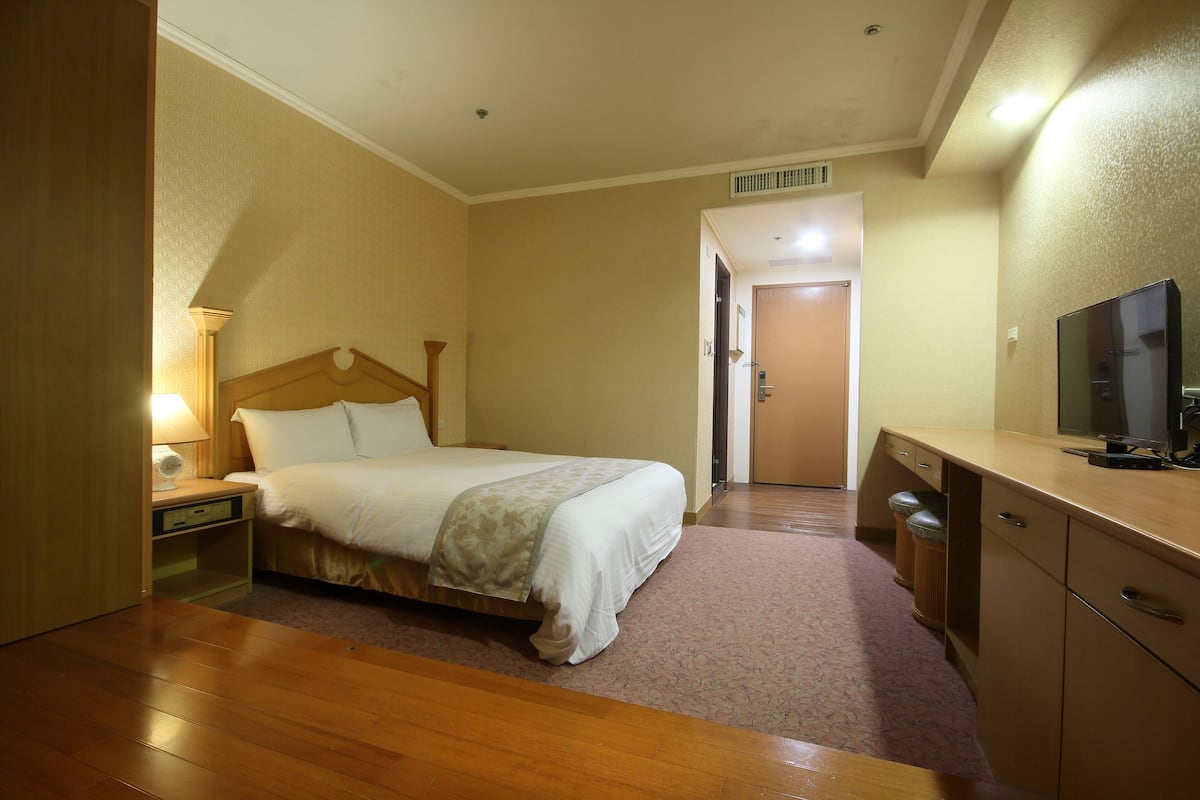 Quarto de hotel com cama, mesa de cabeceira, luminária, televisão e uma porta que dá para outro quarto.
