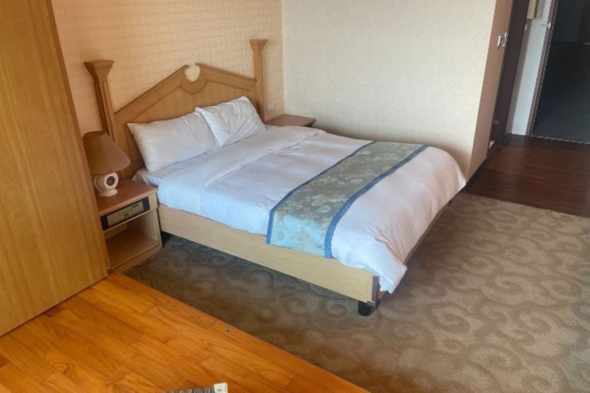 Una cama bien hecha con cabecera de madera, una mesita de noche con lámpara y una alfombra estampada.