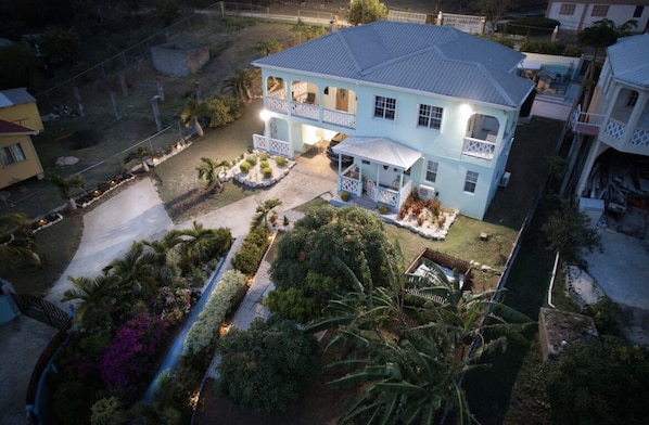Une maison à deux étages dotée d’un balcon, entourée d’un jardin bien entretenu où poussent différentes espèces de plantes, et un chemin mène à l’entrée.