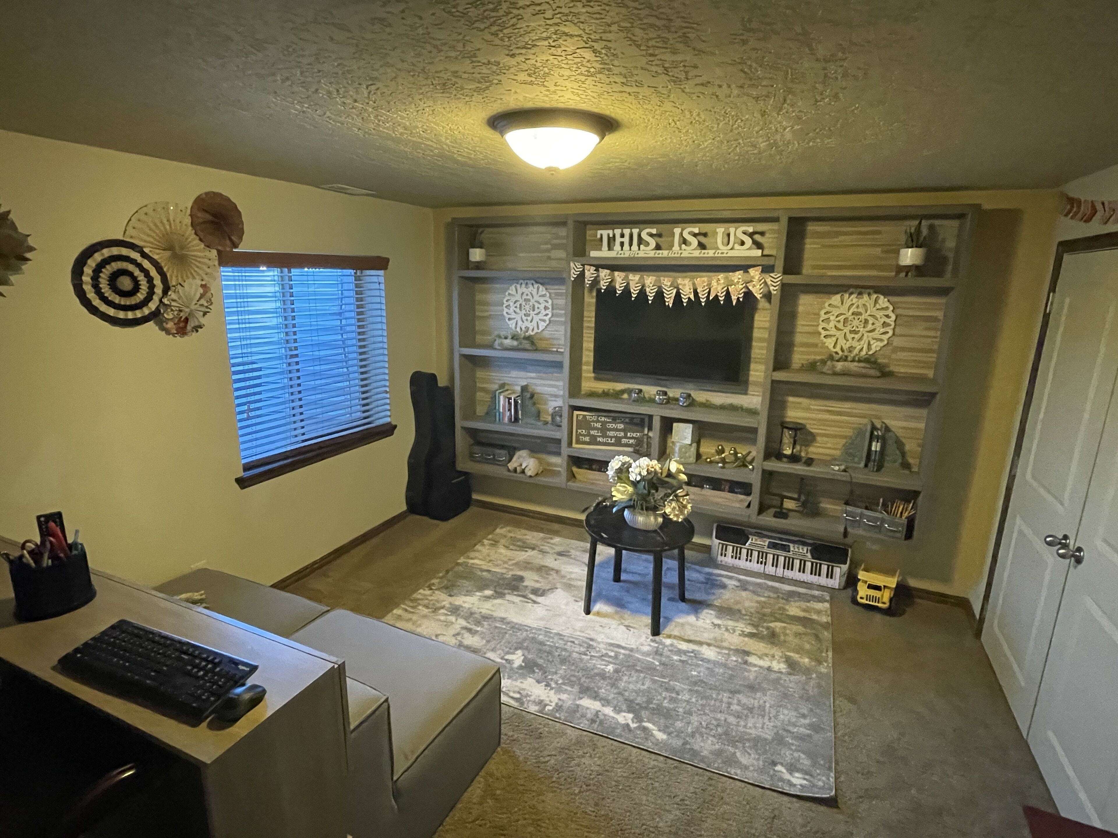 Ein Homeoffice mit Schreibtisch, Tastatur und Maus, einer schwarzen Gitarre, einem Fernseher auf einem Holzregal, Dekoration und einem Teppich.