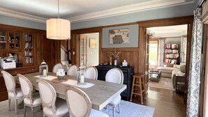Un comedor con una mesa de madera, sillas blancas y una araña. Se observa una estantería con libros y un mapa en la pared.