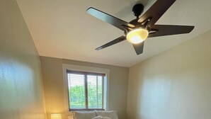 Un dormitorio con cama, mesita de noche, lámpara, ventilador de techo y ventana con persianas.