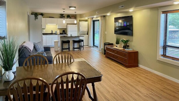 Amplia sala de estar con mesa de comedor de madera, sillas, zona de cocina y televisor de pantalla plana montado en la pared.