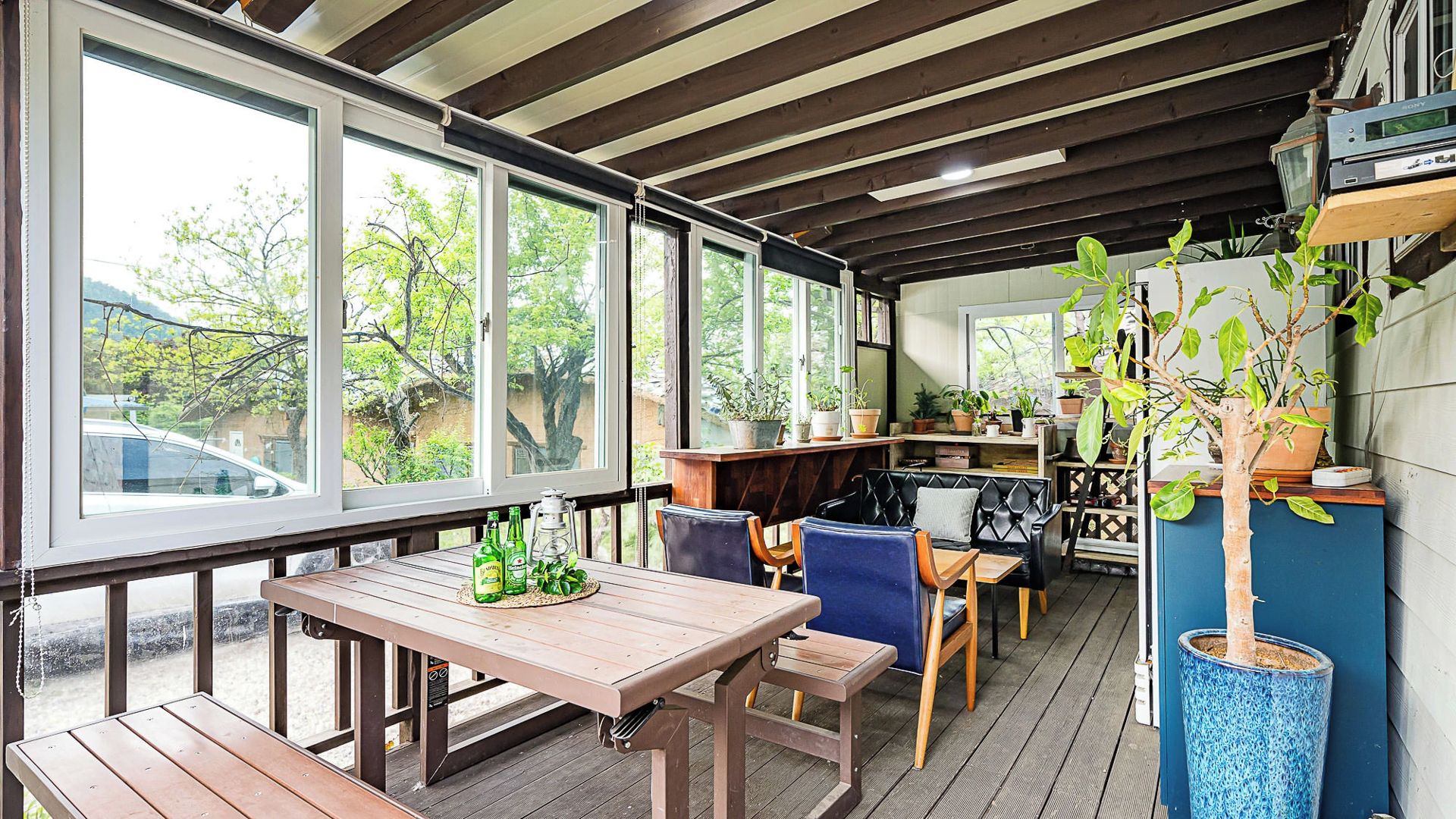 A covered outdoor seating area with wooden tables and chairs, potted plants, and a view of the outdoors.
