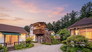A lodge with a wooden staircase leading to a deck, surrounded by lush greenery and a gravel path. - Gapyeong Ojeupension (Gapyeong)