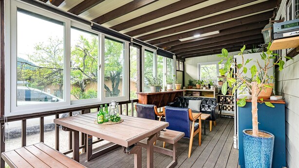 Een overdekt terras met houten tafels en stoelen, planten in potten en uitzicht op de omgeving.