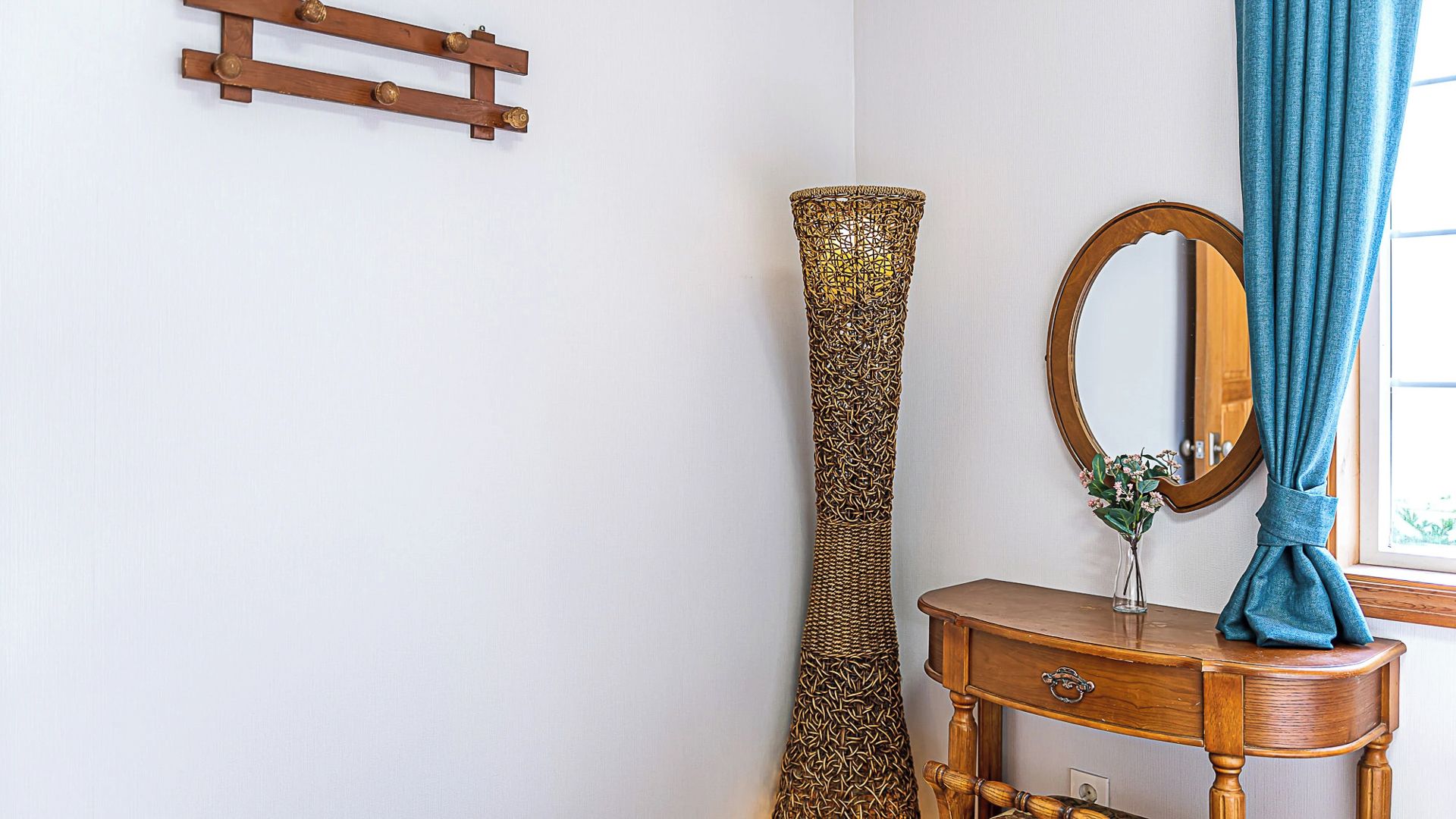 A room with a wooden wall-mounted rack, a tall decorative vase, a round mirror with a wooden frame, and a blue curtain.