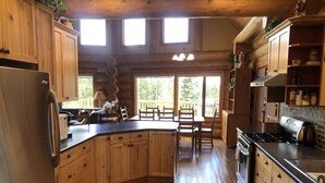 A wooden cabin kitchen with a central island, wooden cabinets, and a dining area with a view of trees. - Cozy Custom Log Cabin Nestled within Mile High Estates (Logan Lake)