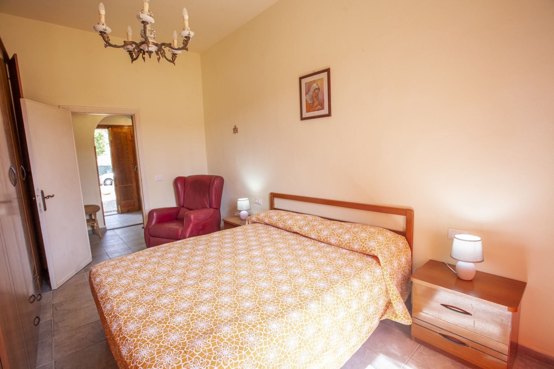 A bedroom with a bed, a chair, a nightstand, a lamp, and a chandelier.