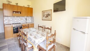 Una cocina funcional con armarios de madera, una mesa de comedor con sillas, un refrigerador y un televisor instalado en la pared.