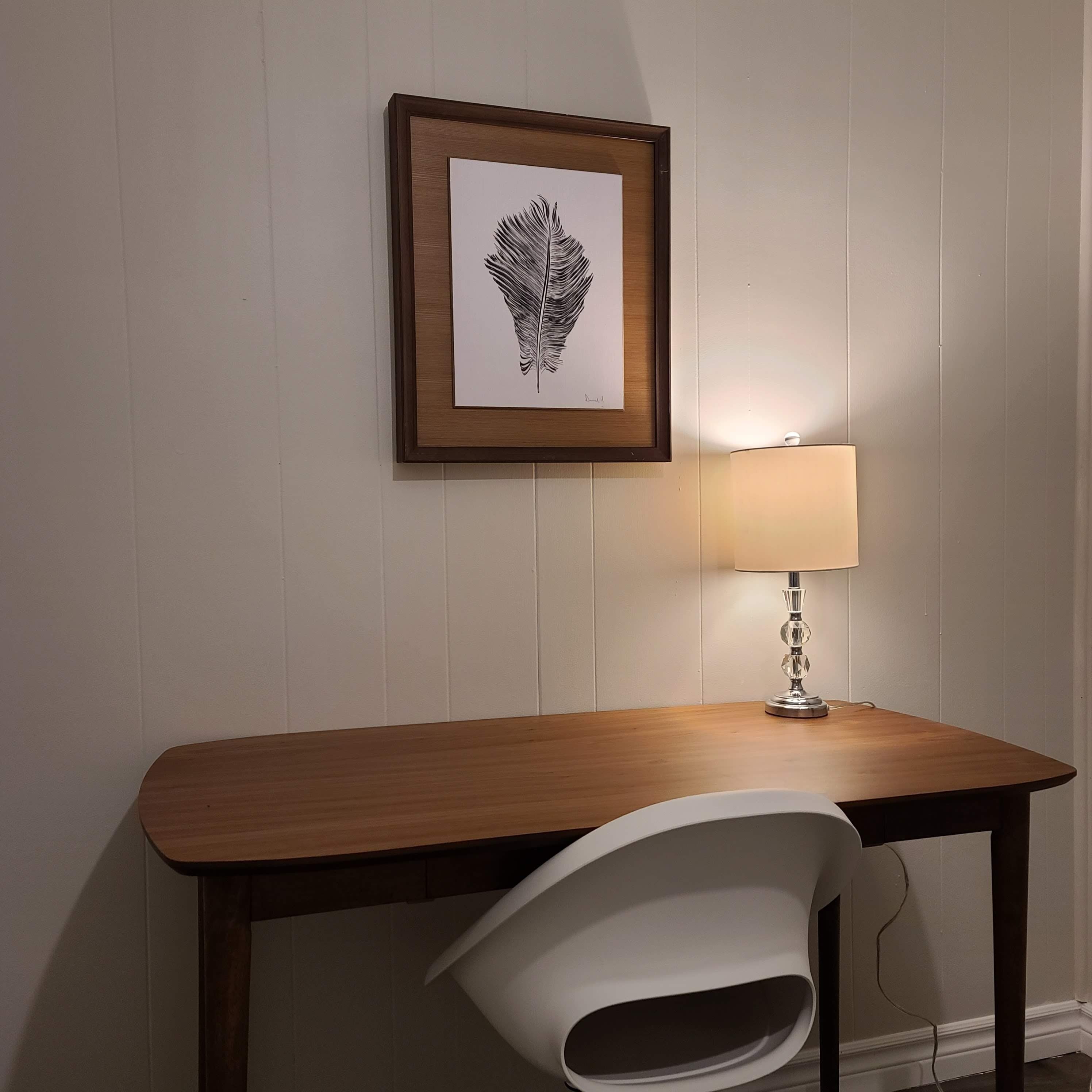 A wooden desk with a white chair, a framed artwork of a feather, and a table lamp.