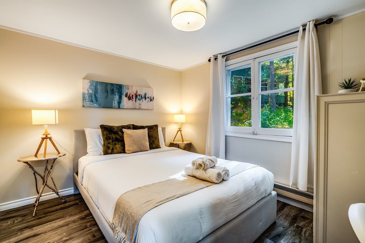 Ein ordentlich gemachtes Bett mit weißen Leinentüchern und einem zusammengerollten Handtuch, zwei Nachttischlampen, ein Bild an der Wand und ein Fenster mit Vorhängen.