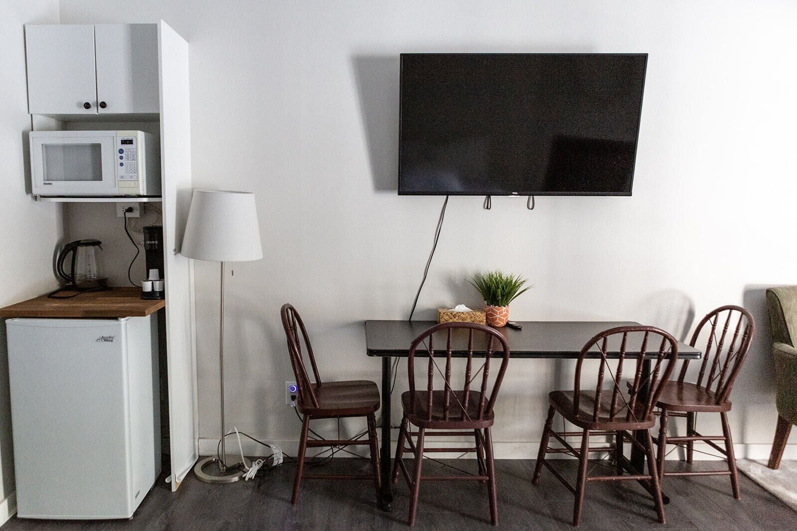 A compact room with a dining table, four chairs, a flat-screen TV mounted on the wall, a microwave, a coffee maker, and a small refrigerator.