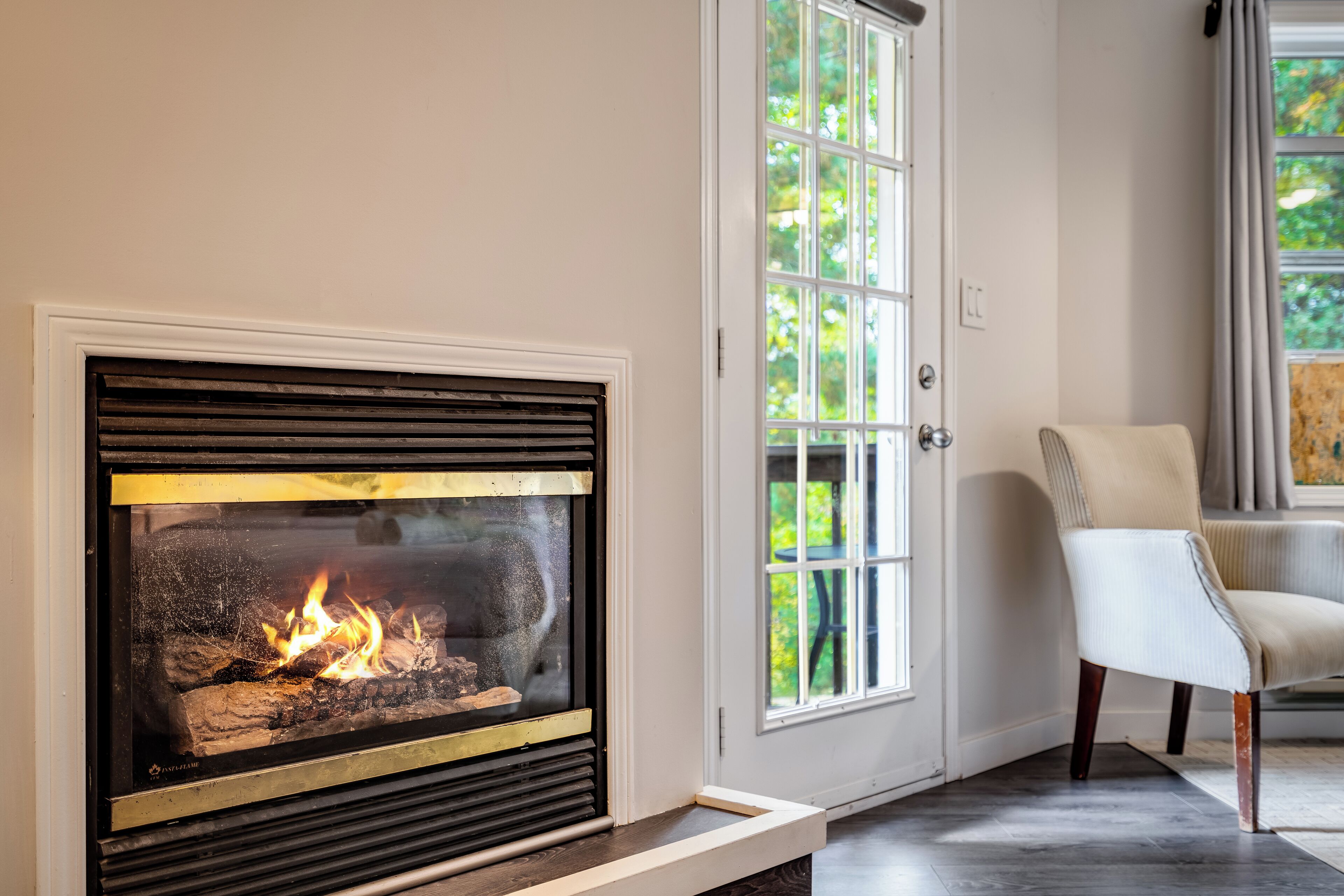 Une cheminée moderne avec une façade en verre, une porte blanche dotée de panneaux de verre, et un fauteuil dans une pièce au sol carrelé.