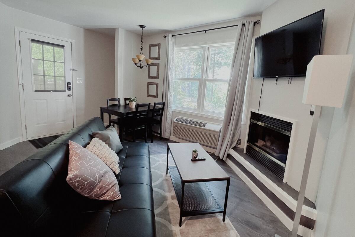 Ein Wohnzimmer mit schwarzem Ledersofa, einem Esstisch mit Stühlen, einem an der Wand montierten Fernseher und einem Kamin.
