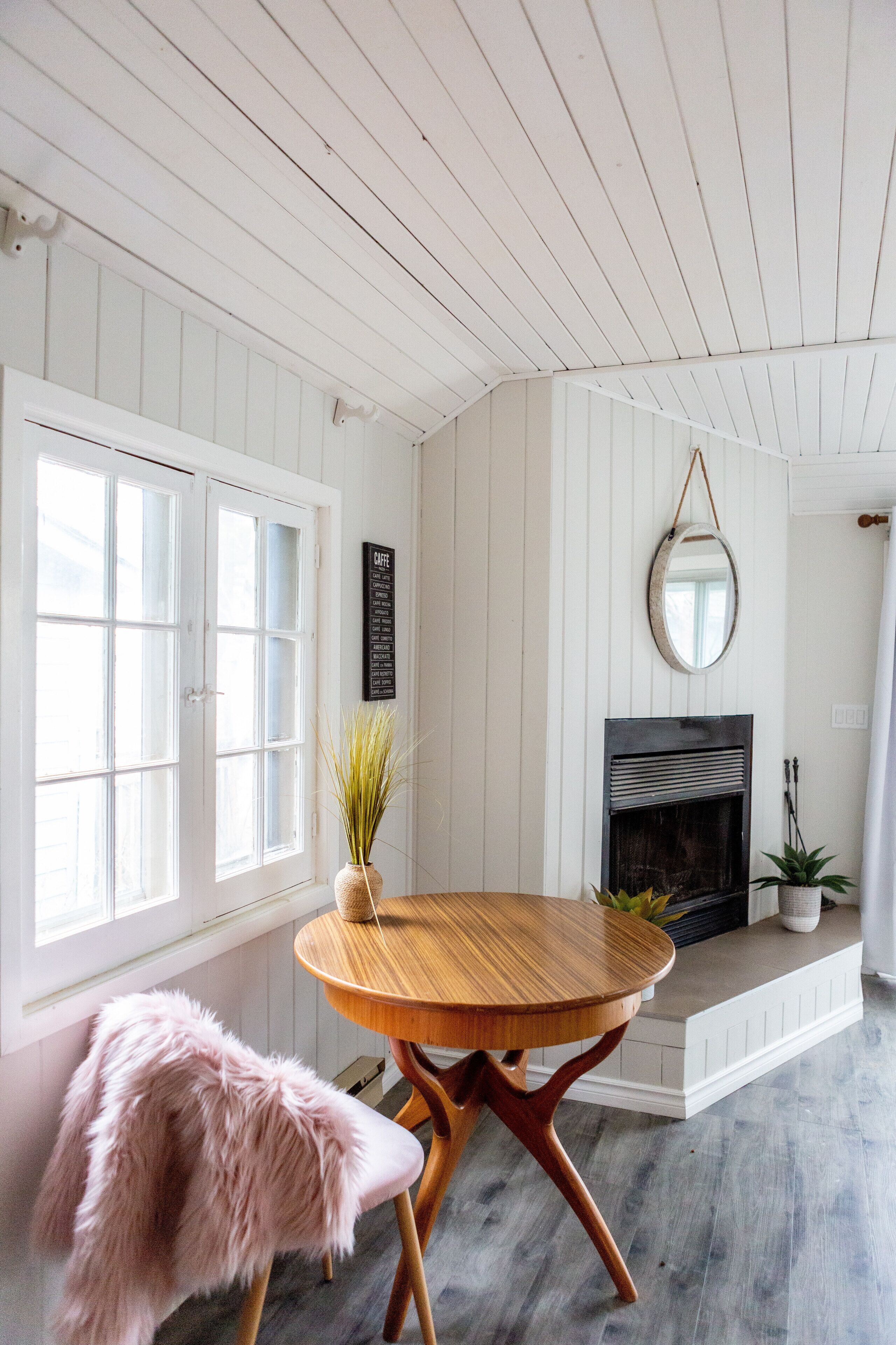 A room with a wooden dining table, a pink chair, a fireplace, and a window with white shutters.