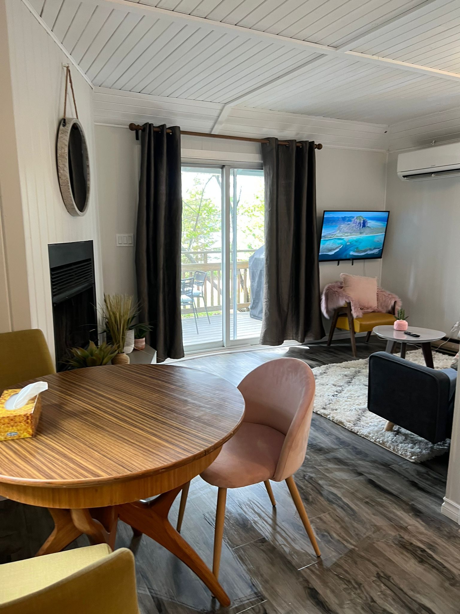 A modern living room with a wooden dining table, two chairs, a fireplace, a television, and a view of the outdoors through a sliding glass door.
