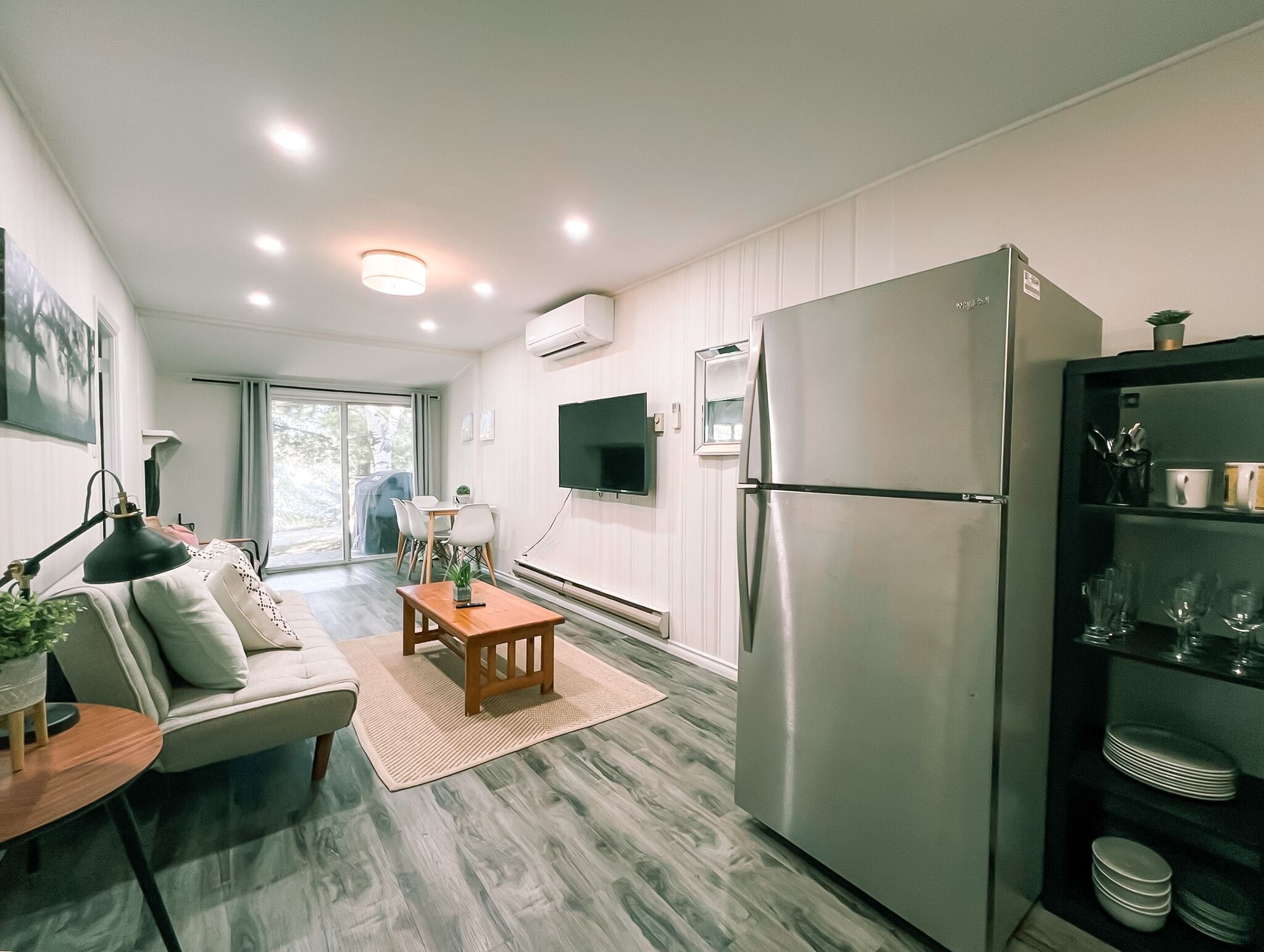 A modern kitchen with a stainless steel refrigerator, a dining area with a sofa and table, and a TV mounted on the wall.