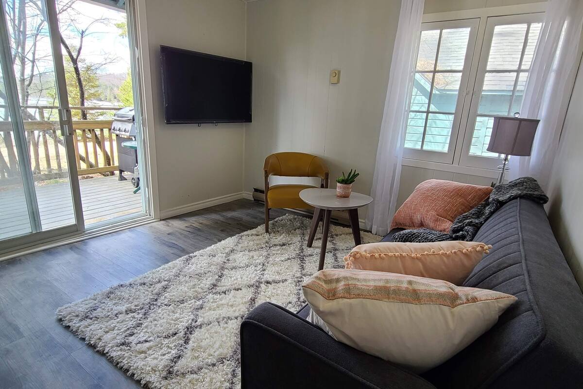 Ein Wohnzimmer mit einem dunklen Sofa, einem kleinen runden Tisch, einem Flachbildfernseher und einem großen Fenster mit Vorhängen.