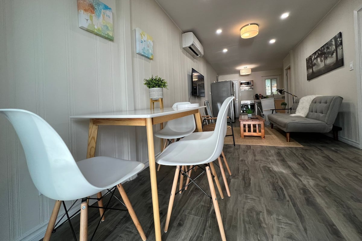 Ein modernes Wohnzimmer mit Essbereich, ausgestattet mit einem grauen Sofa, weißen Stühlen, einem Holztisch und einem grauen Teppich.