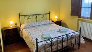 A bedroom with a bed, two bedside tables with lamps, a window with wooden shutters, and a radiator.