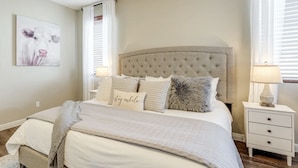 A bedroom with a tufted headboard, a bed with multiple pillows, a bedside table with a lamp, and a wall-mounted artwork of a cow. - Young Desert Oasis with Heated Pool in Estrella Mountain Ranch (Goodyear)