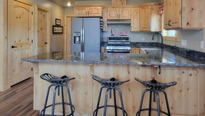A kitchen with wooden cabinets, a granite countertop, a stainless steel refrigerator, a stove, and three bar stools. - Welcome to Duck Creek Haven A fully furnished cabin secluded in a serene setting (Duck Creek Village)