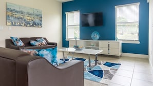 Un salon avec un mur bleu, un téléviseur à écran plat, un canapé avec des coussins bleus, une table basse et un tapis décoratif.