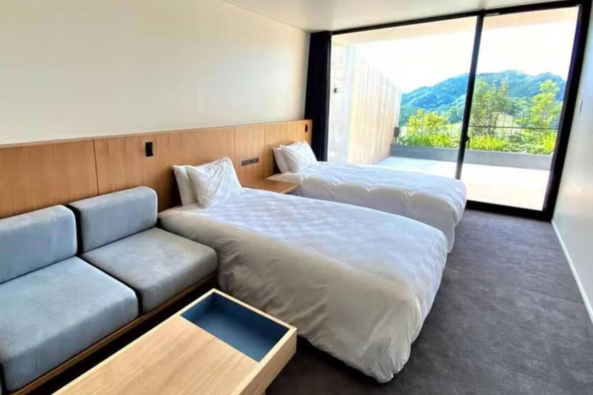 A hotel room with a sofa, two beds, a small table, and a large window with a view of greenery.