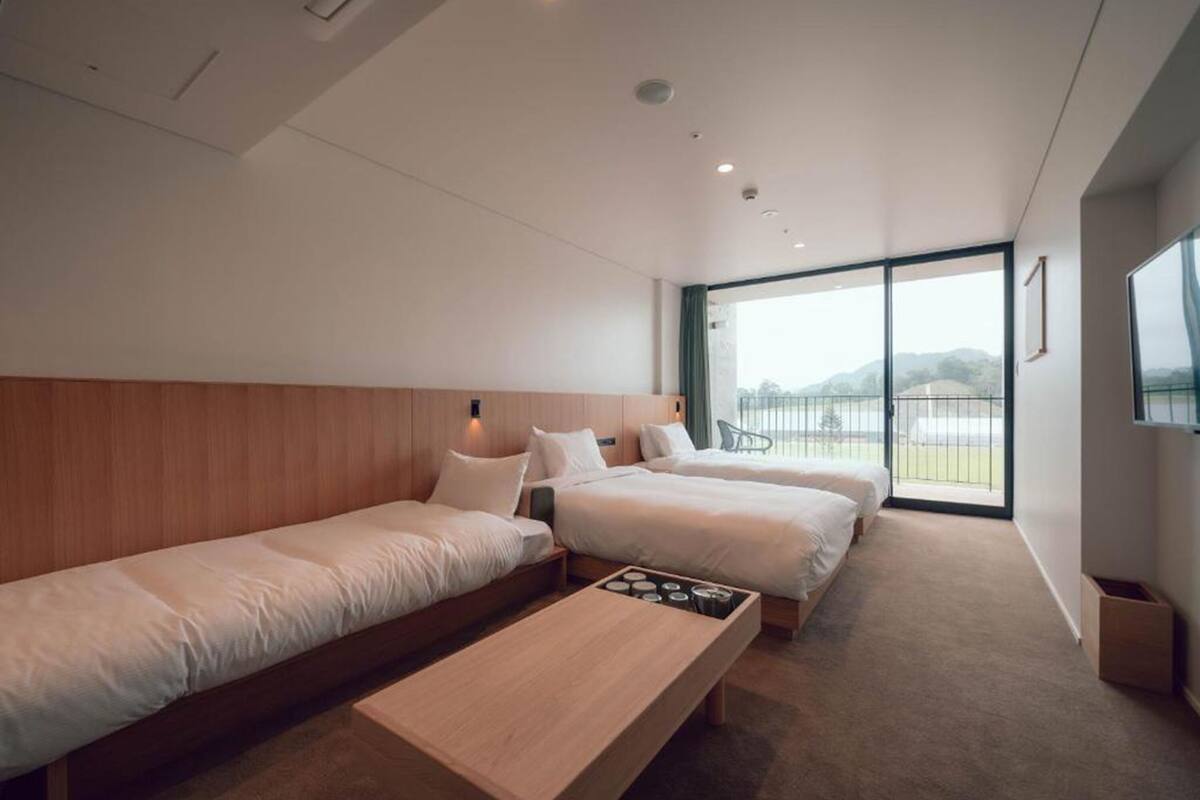 A modern hotel room with two single beds, a wooden bench, a small table, and a large window offering a view of the outdoors.