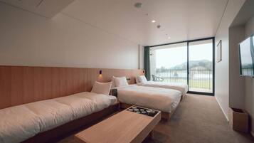 Quarto de hotel moderno com duas camas de solteiro, um banco de madeira, uma mesinha e uma janela ampla com vista para o exterior.